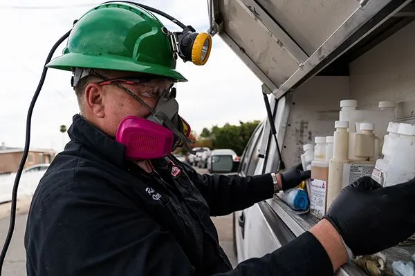 Pest control pro with hardhat and face mask gathers chemical bottles from work truck